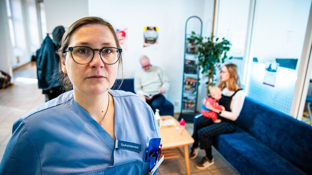 En dyster Jessica Eek iklädd sjuksvårdskläder i ett väntrum på sjukhset